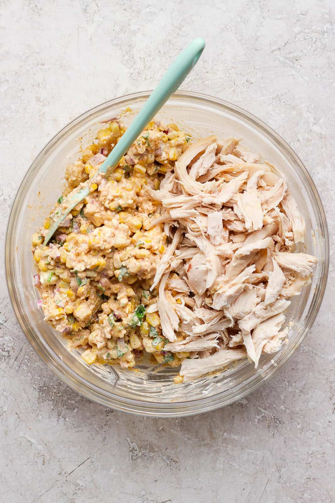 Cooked, shredded chicken added to the Mexican street corn mixture in a glass bowl with a spatula sticking out the side.