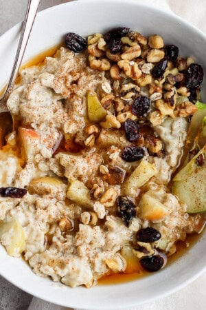 Apple pie oatmeal in a bowl topped with apple slices, raisins, and maple syrup.