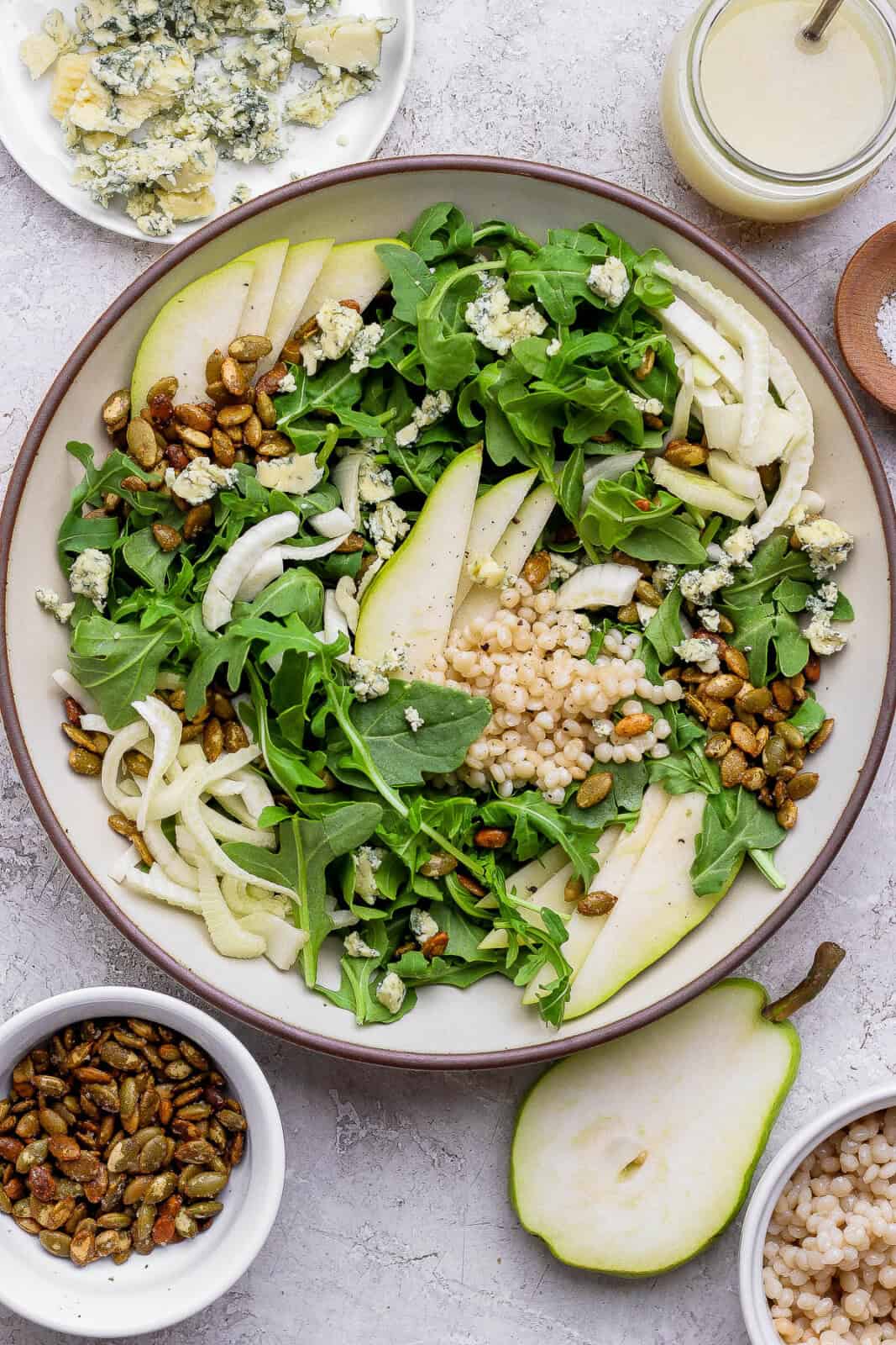 Fennel pear salad in a large bowl next to small bowls of pepitas, fennel, dressing, and half a pear.