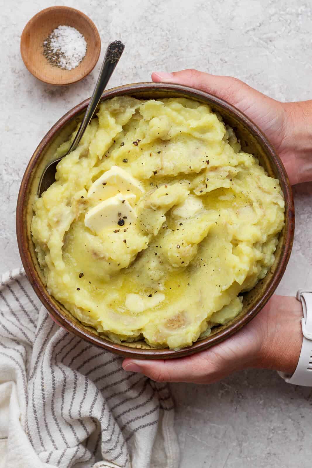 Two hands holding a bowl of horseradish mashed potatoes next to a small dish of salt and pepper.