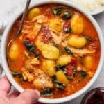 A hand holding a bowl of marry me chicken gnocchi soup with a spoon sticking out the side.