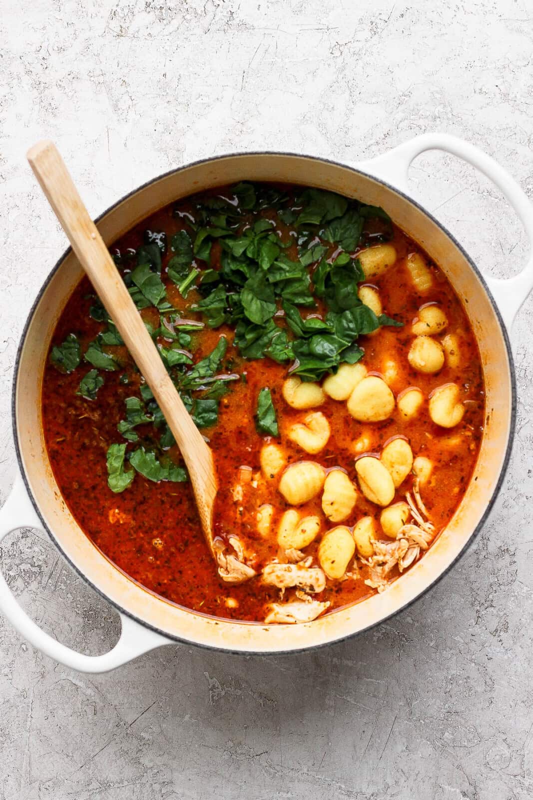 Shredded chicken, gnocchi, and fresh spinach added to a pot of marry me chicken gnocchi soup with a wooden spoon sticking out the side.