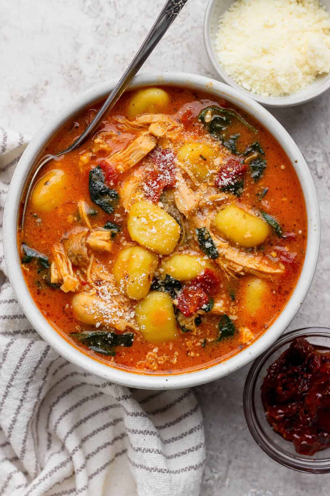 A bowl of marry me chicken gnocchi soup next to a kitchen towel and small bowls of parmesan and sun-dried tomatoes.