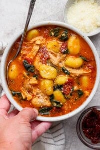 Someone holding a bowl of marry me chicken gnocchi soup next to small bowls of parmesan cheese and sun-dried tomatoes.