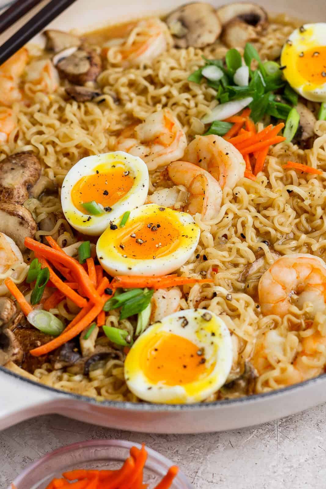 A soft boiled egg cut in half and placed on a skillet of shrimp ramen noodles with some carrots and sliced green onions.
