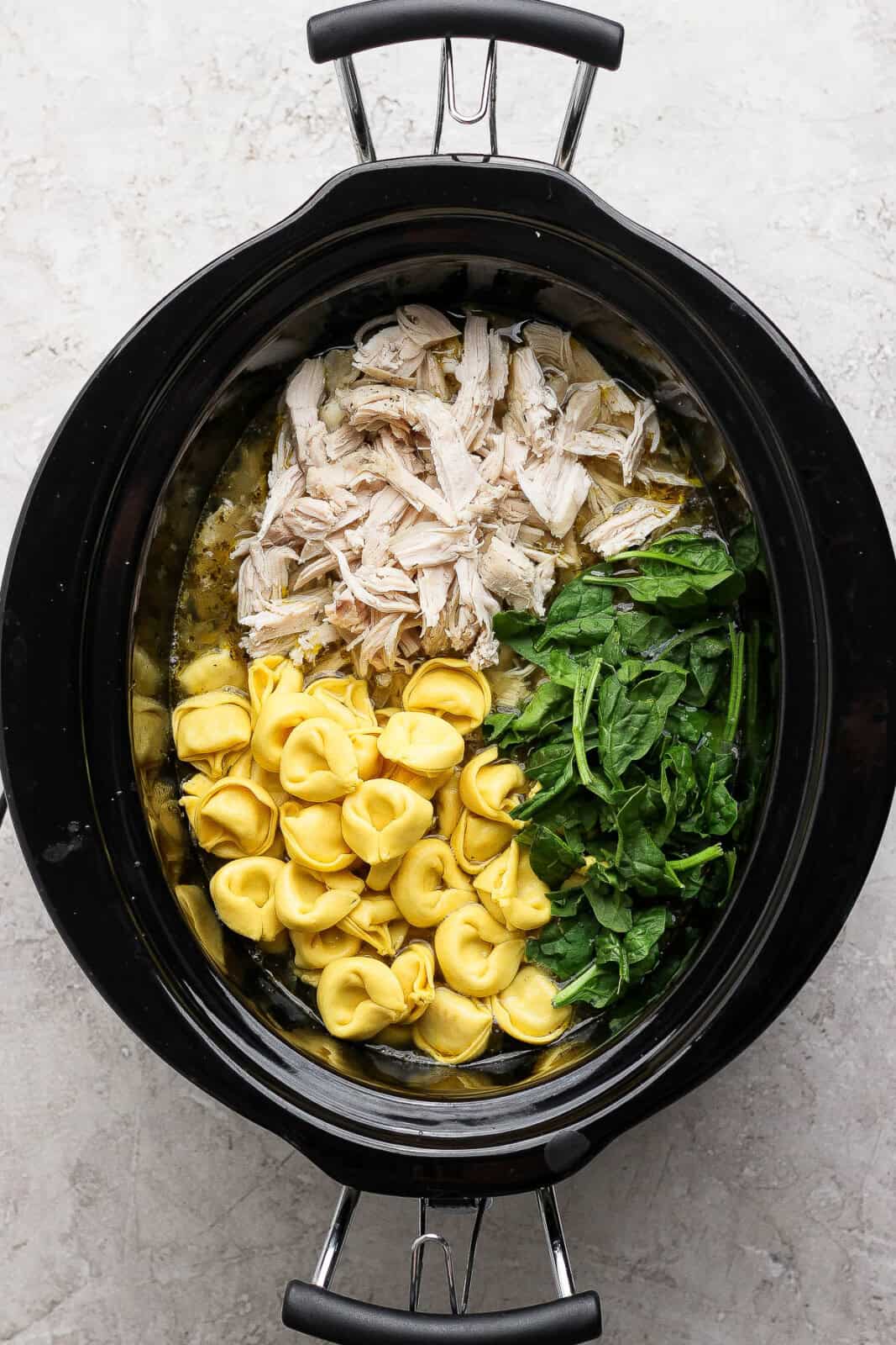 Shredded chicken, cheese tortellini, and spinach added to the slow cooker to make slow cooker chicken tortellini soup.