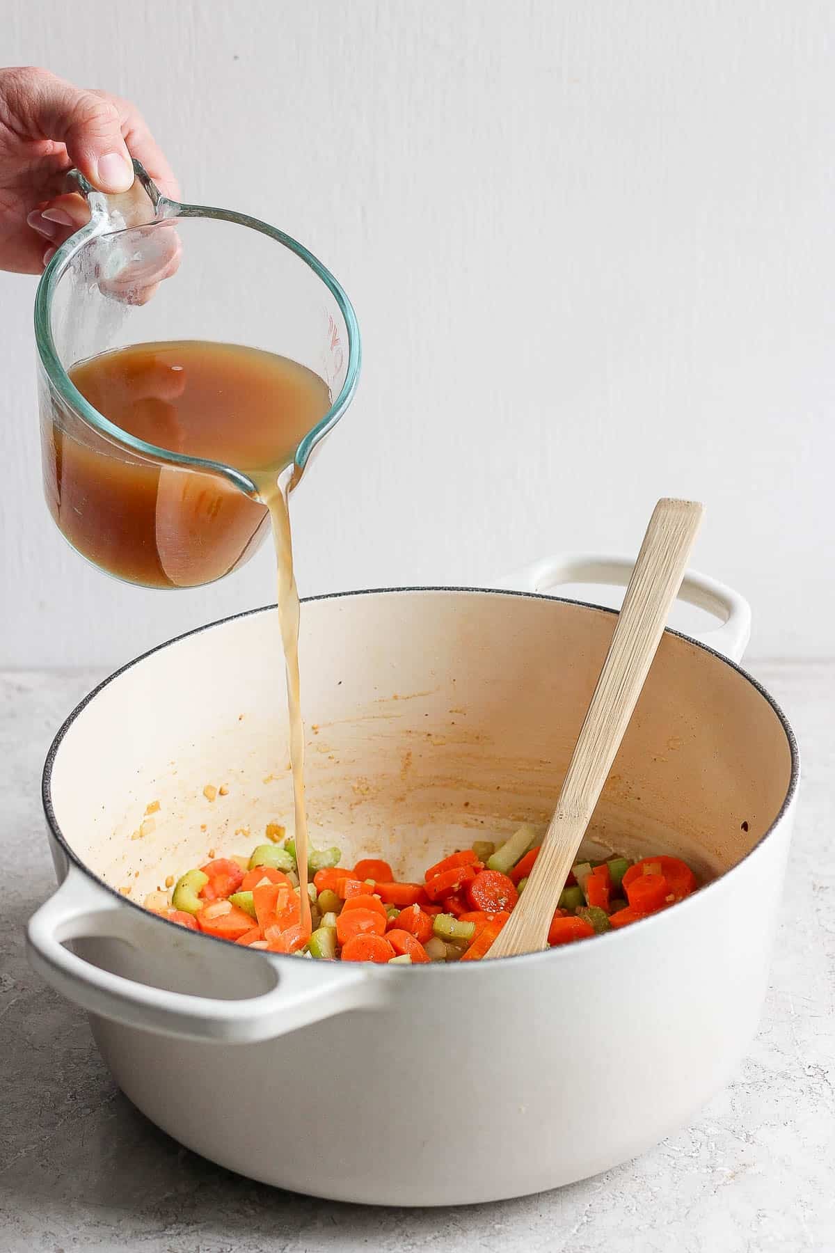 A hand pouring chicken bone broth into dutch oven to make ginger turmeric chicken soup.